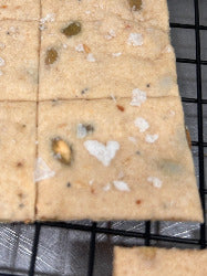 A close-up of a rectangular beige baked good, likely a flatbread or cracker, topped with coarse white salt flakes and small green and brown inclusions, resting on a black wire cooling rack.